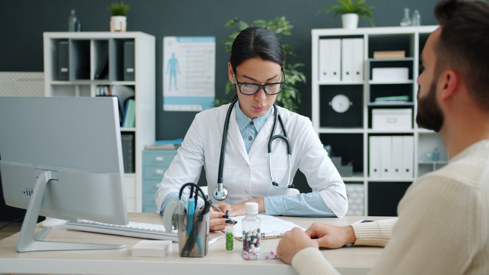 A clinician working at a desk with a computer, medical files and records in the office behind her